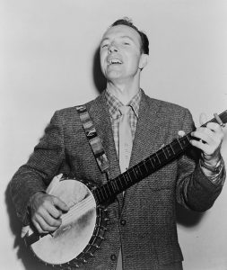 Black and white photographic image of Pete Seeger playing a banjo and singing.