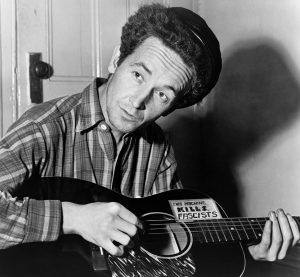 Black and white photographic image of Woody Guthrie playing a guitar. The sticker on the guitar reads "This machine kills fascists"