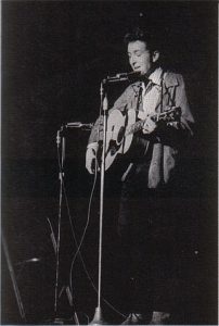 Black and white photographic image of Bob Dylan playing a guitar and singing into a microphone. It appears that he might be performing.