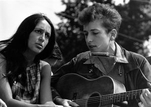 Black and white photographic image of Bob Dylan playing a guitar and singing into a harmonica with Joan Baez.