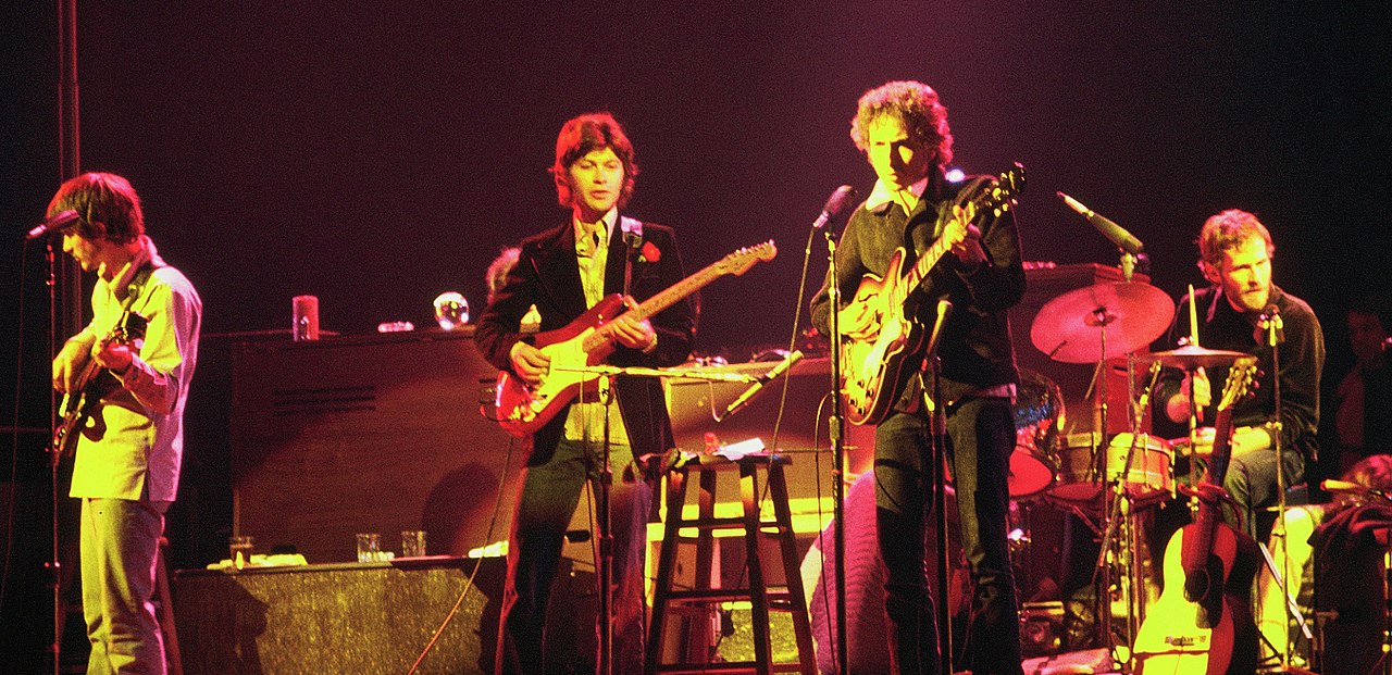 Color image of Bob Dylan and The Band. There are three guitarists and one drummer pictured. The picture has a red-yellow glow to it.