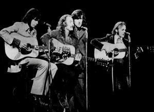 Black and white photographic image of Crosby, Stills, and Nash performing. There are four men in this photo. Three of the four have guitars.