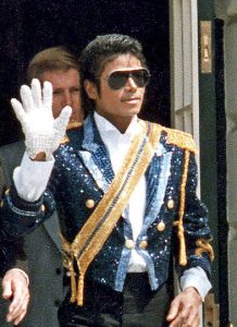 Photographic image of Michael Jackson holding up his hand wearing a sparkly suit, white gloves, a yellow sash, and sunglasses.