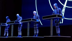 Color photo of Kraftwerk live dressed up with blue-purple lighting highlighting the white lines on their outfits.
