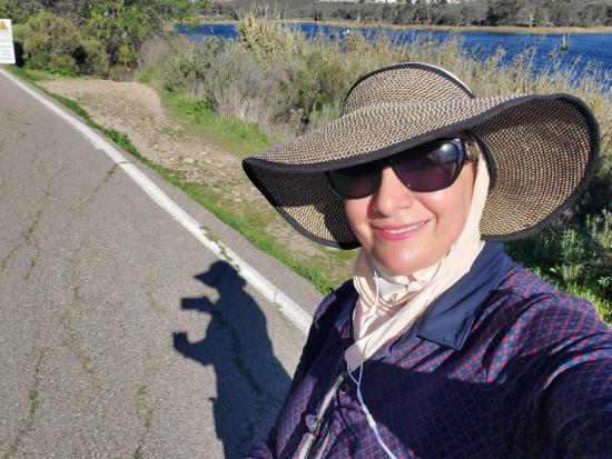 A woman is wearing a hat and sunglasses. She is standing on the side of the road and is taking a selfie. Her shadow is in the picture.