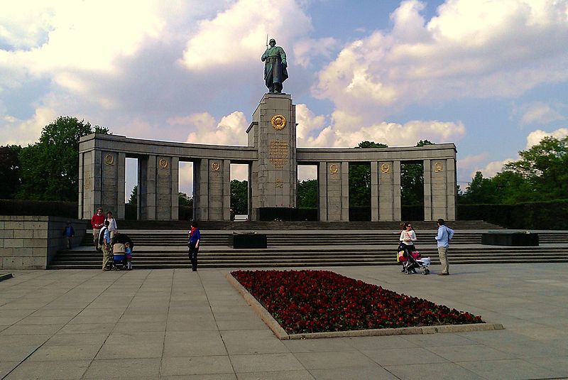 A photo of the Soviet War Memorial in Berlin. A large bed of red roses lays in the middle of the ground. In the background a monument stands with a statue of an person on top.